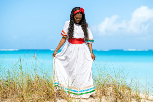 Woman wearing a white dress with a red sash in a sea oat dune by the beach.