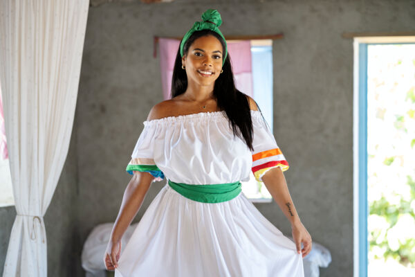 Woman wearing a white dress and green sash inside a traditional home. 