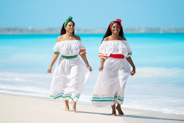 Women in white dresses with colorful sashes walking along a Turks and Caicos beach.