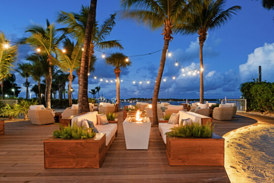 Upscale lounge area with fire tables, palm trees, and string lights