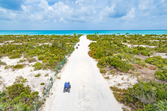 ATVs driving down a dirt path to a beautiful white-sand beach with turquoise water. 