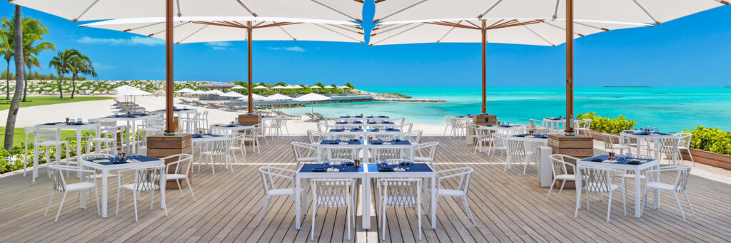 Dining tables and umbrellas at a waterfront restaurant overlooking a turquoise ocean.